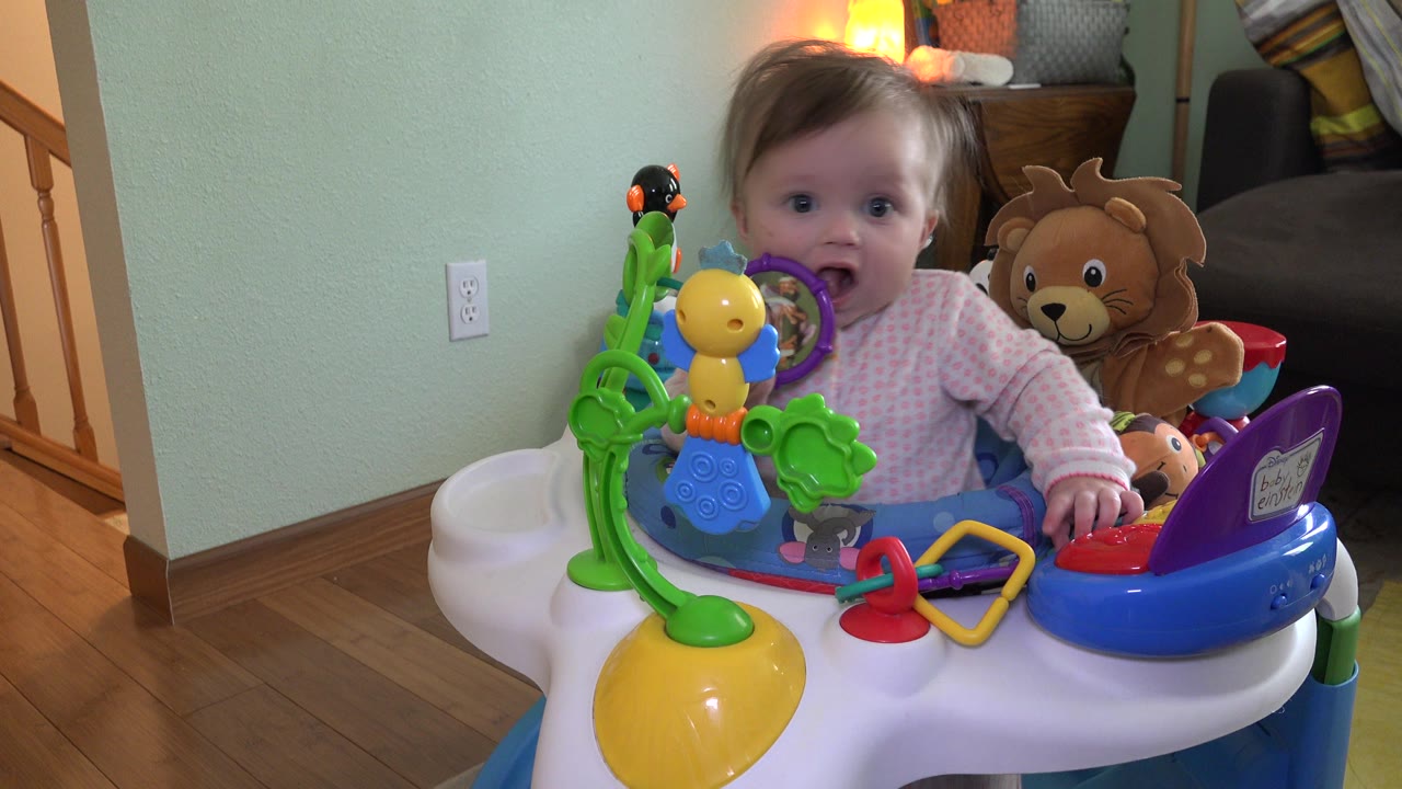 Toddler remains front-facing in the Westminster home interior, framed by a colorful activity chair and plush animal toys in the background.