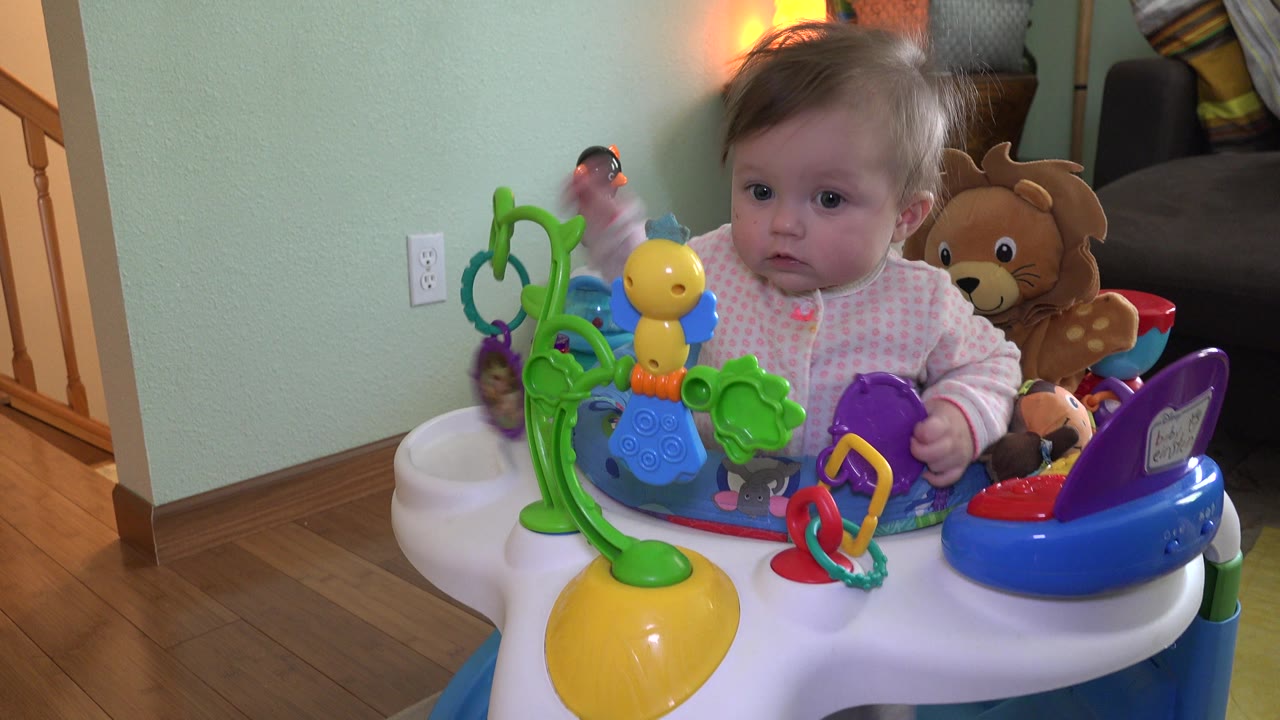 At 0:02, the baby girl faces forward from the walker, with a yellow toy, green play arch, purple tray, stuffed lion, and warm lamp in the background.