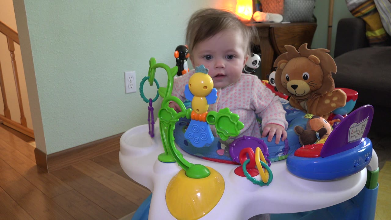 Baby sitting in a colorful activity seat with toys indoors.