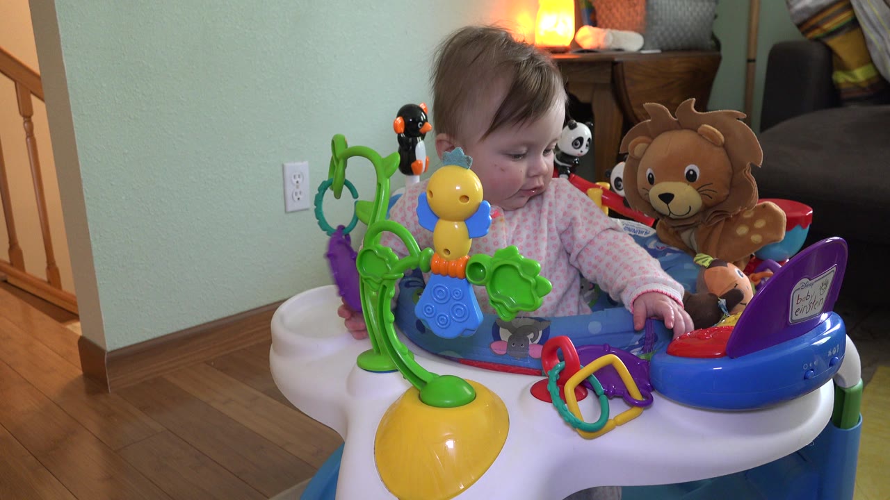 Baby sits in a colorful activity saucer in a home interior.