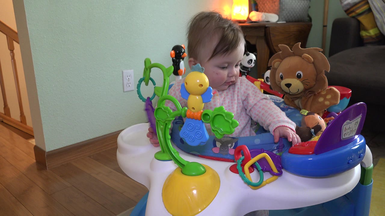 At 0:01, the baby looks down toward the toy tray in the Westminster home, with a yellow hanging toy, purple panel, and plush lion around the seat.