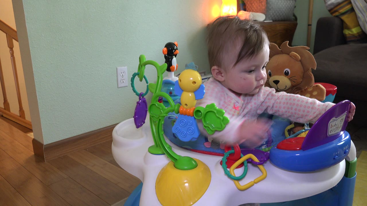Baby girl reaches across a colorful activity bouncer in a home interior.