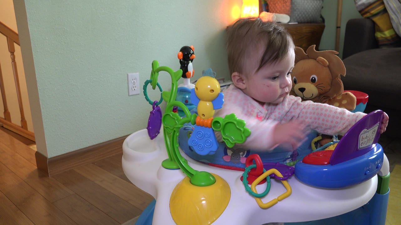 At 0:01 in Westminster, Colorado, the baby girl leans forward in the bouncer as her hand blurs near the red and purple toy controls.