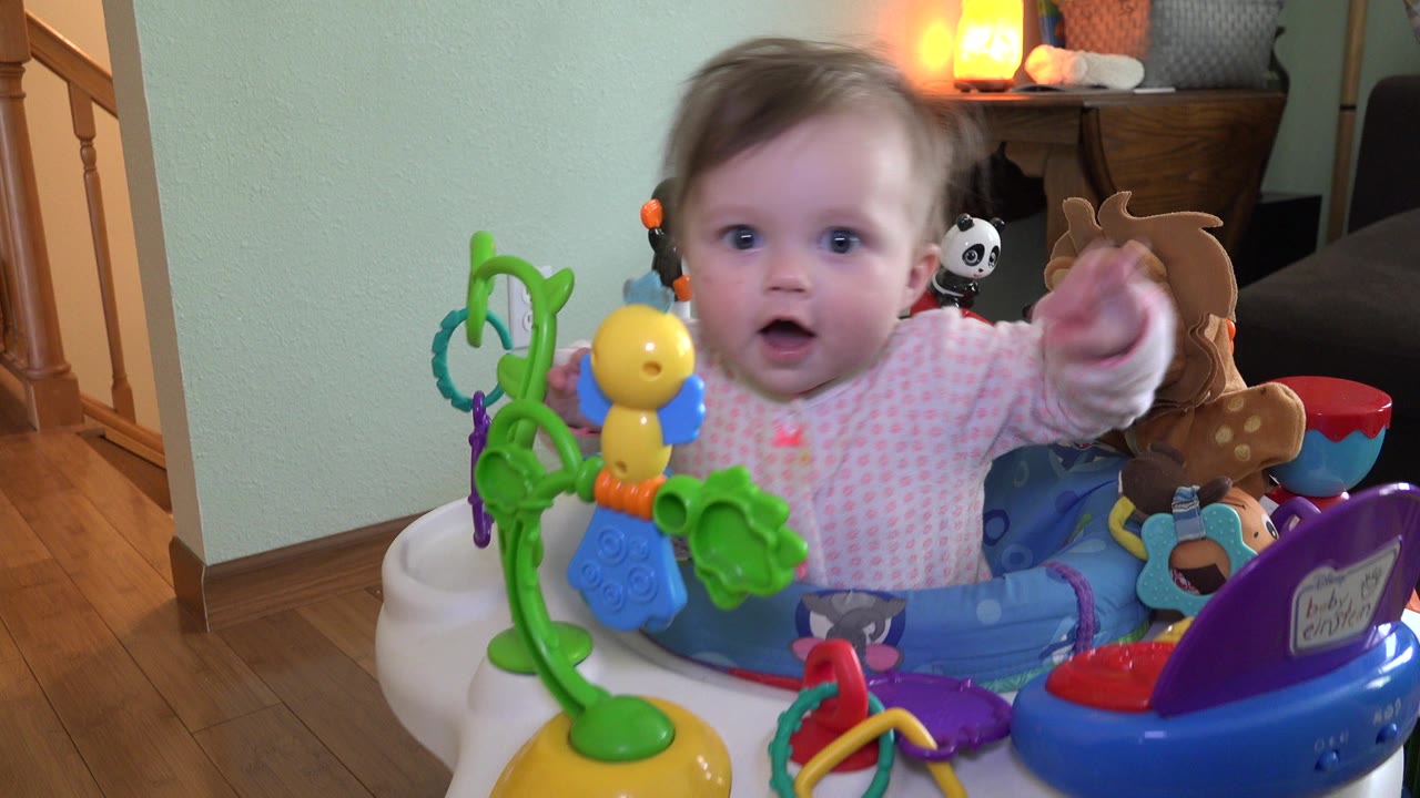 At 0:01, the baby faces forward with an arm extended from the blue activity jumper, surrounded by bright plastic toys and stuffed animals in the Westminster, Colorado interior.