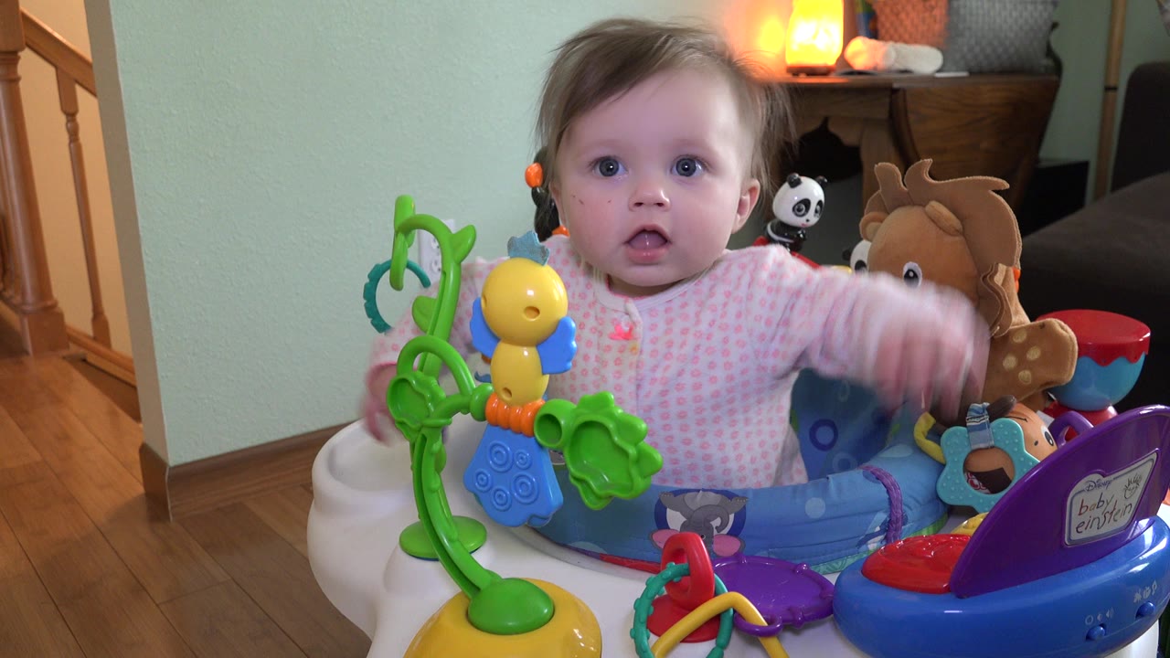 A toddler in Westminster, Colorado sits centered in a blue activity bouncer with green and yellow toys in front and warm lamp light behind.