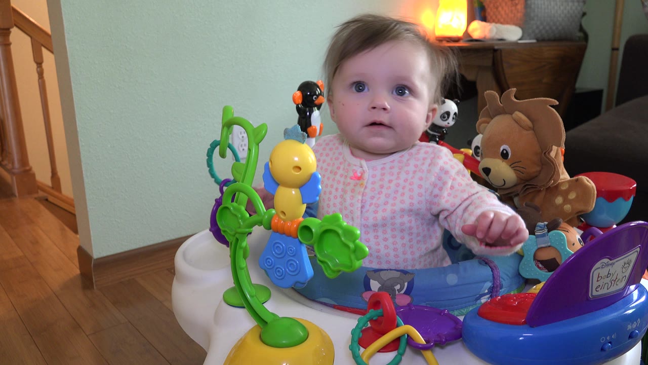 A toddler in Westminster, Colorado looks forward from the bouncer with a green toy arch and yellow toy bird prominent at the front.