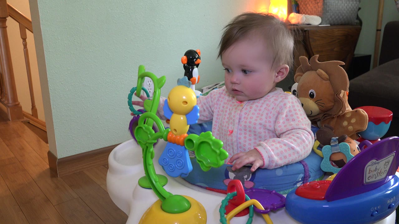 At 0:00, a baby in a pink outfit sits in a blue activity walker in a Westminster, Colorado home, looking left toward green and yellow toys with wood flooring and stairs behind.