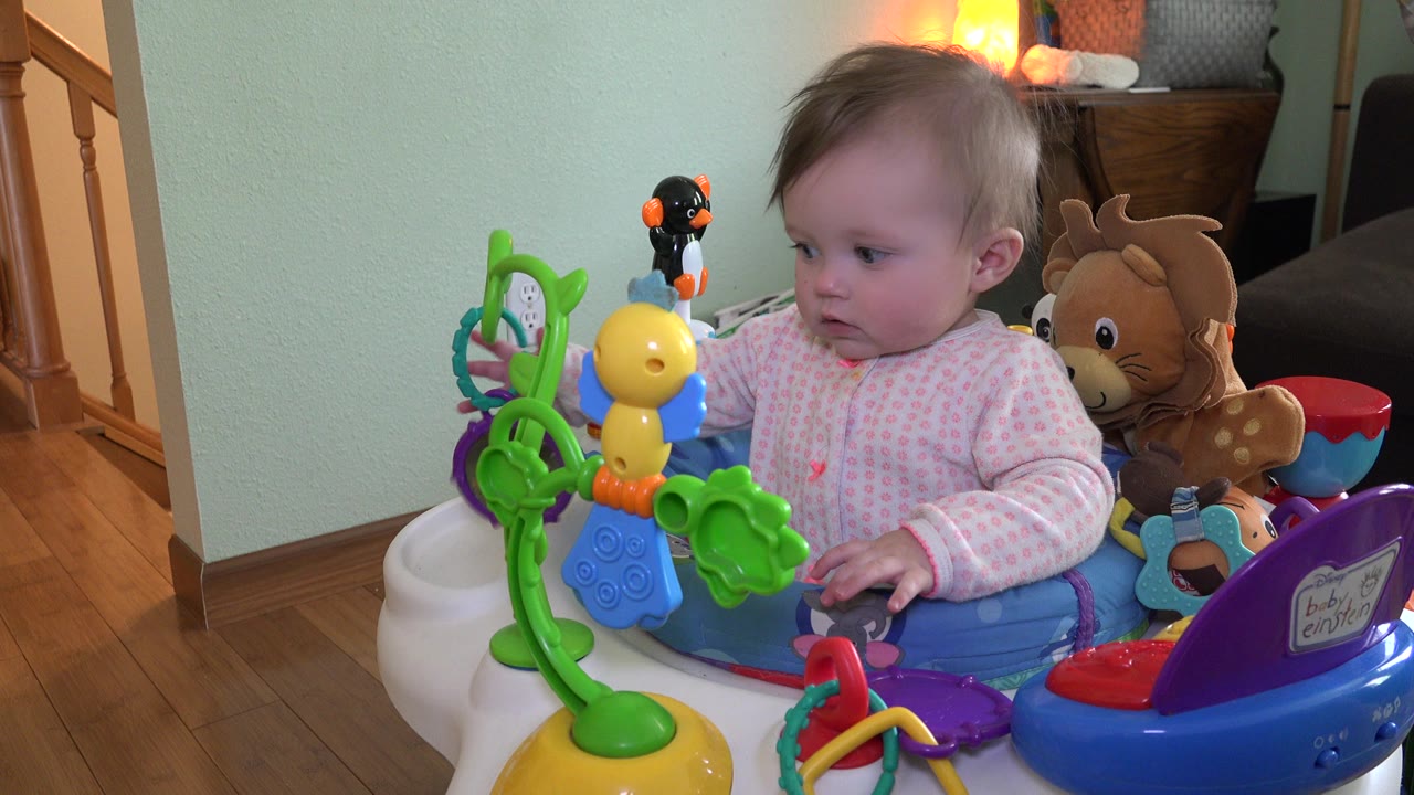 At 0:01, the baby remains turned left in the activity walker, with the toy cluster filling the foreground and a plush deer, lamp, and stair rail visible in the Westminster home interior.
