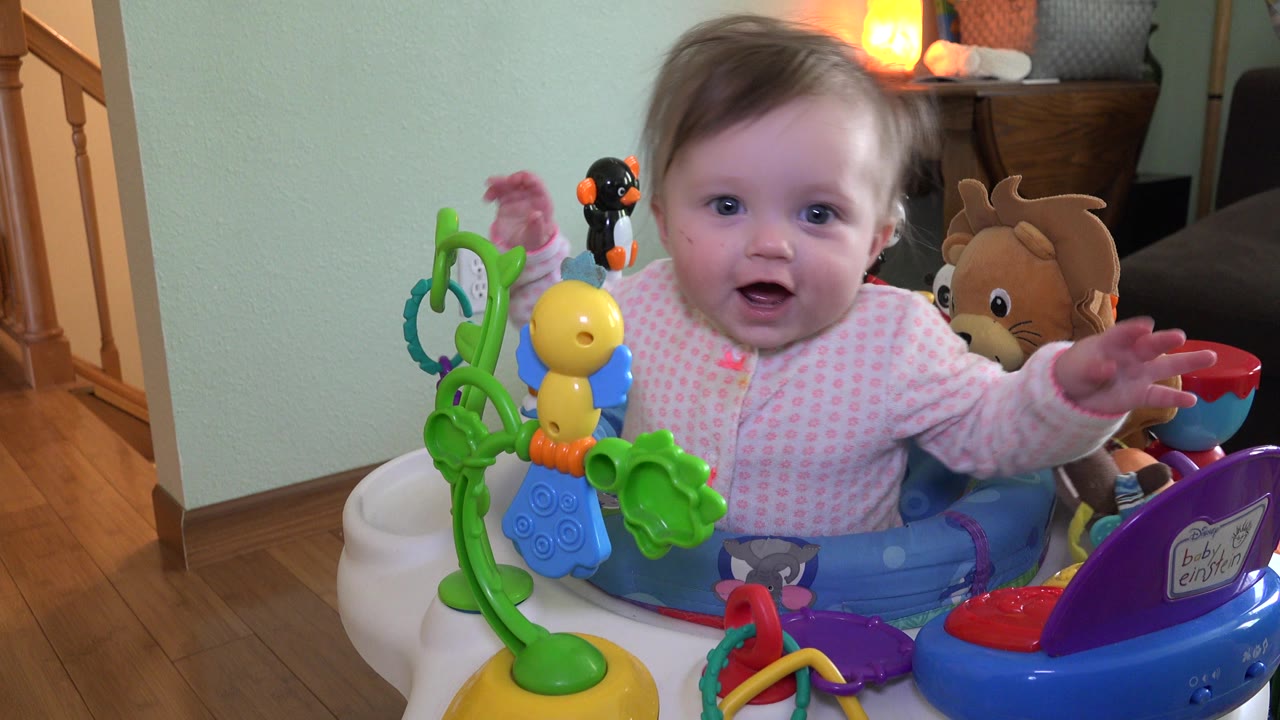 Baby in pink clothes sits in a blue activity seat surrounded by colorful toys.