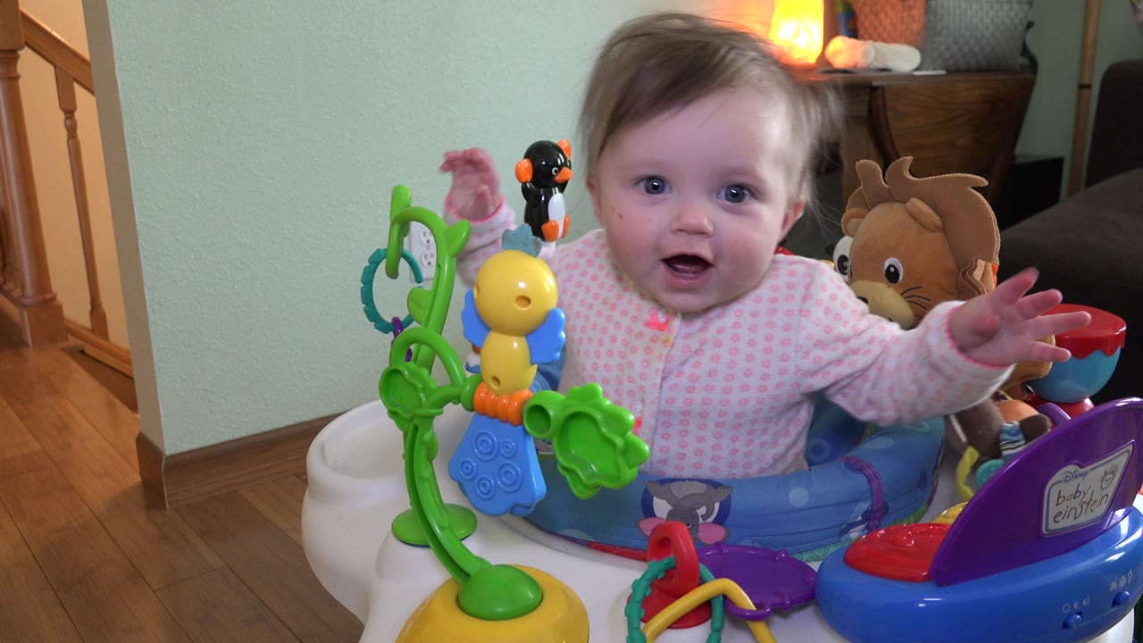 In Westminster, Colorado, the baby raises a hand toward the camera while seated behind colorful activity toys, with a staircase and warm lamp visible behind.