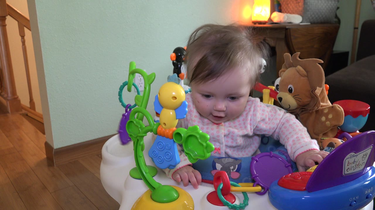 At 0:00, a baby in pink clothing sits in a colorful toy activity seat inside a Westminster, Colorado home, with stairs and warm lamp light in the background.