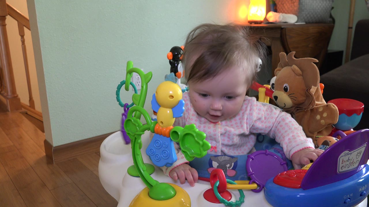 At 0:01, the baby leans forward toward the toy tray, with green, yellow, blue, purple, and red play pieces filling the foreground of the Westminster home scene.