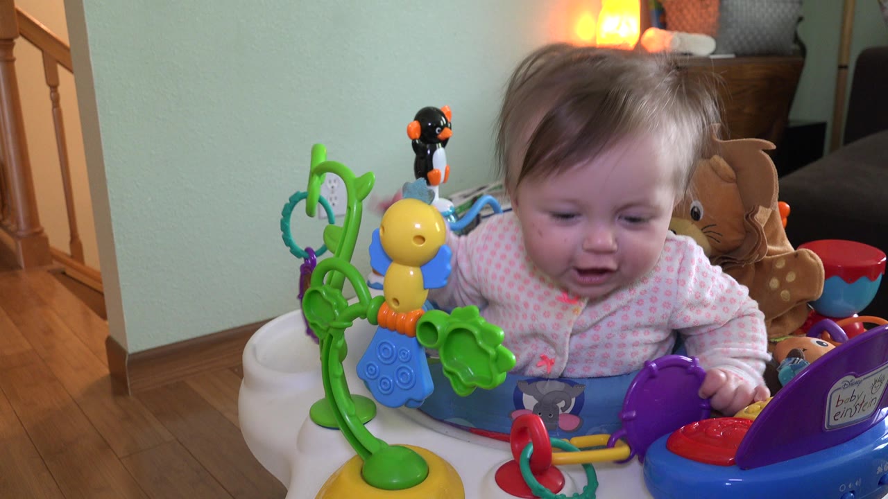 The baby remains in the Westminster home toy seat, looking down toward the purple and red activity pieces while plush toys and a glowing lamp sit behind.