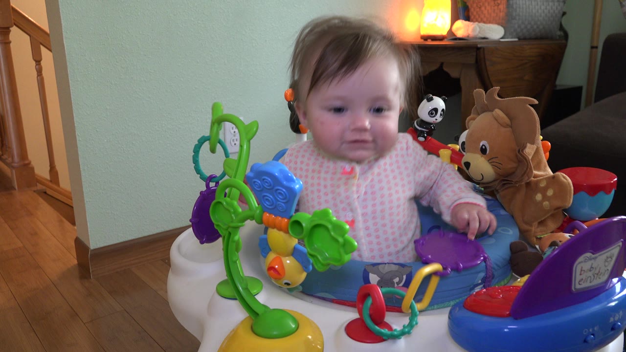 Baby in pink pajamas sitting in a colorful activity seat indoors.