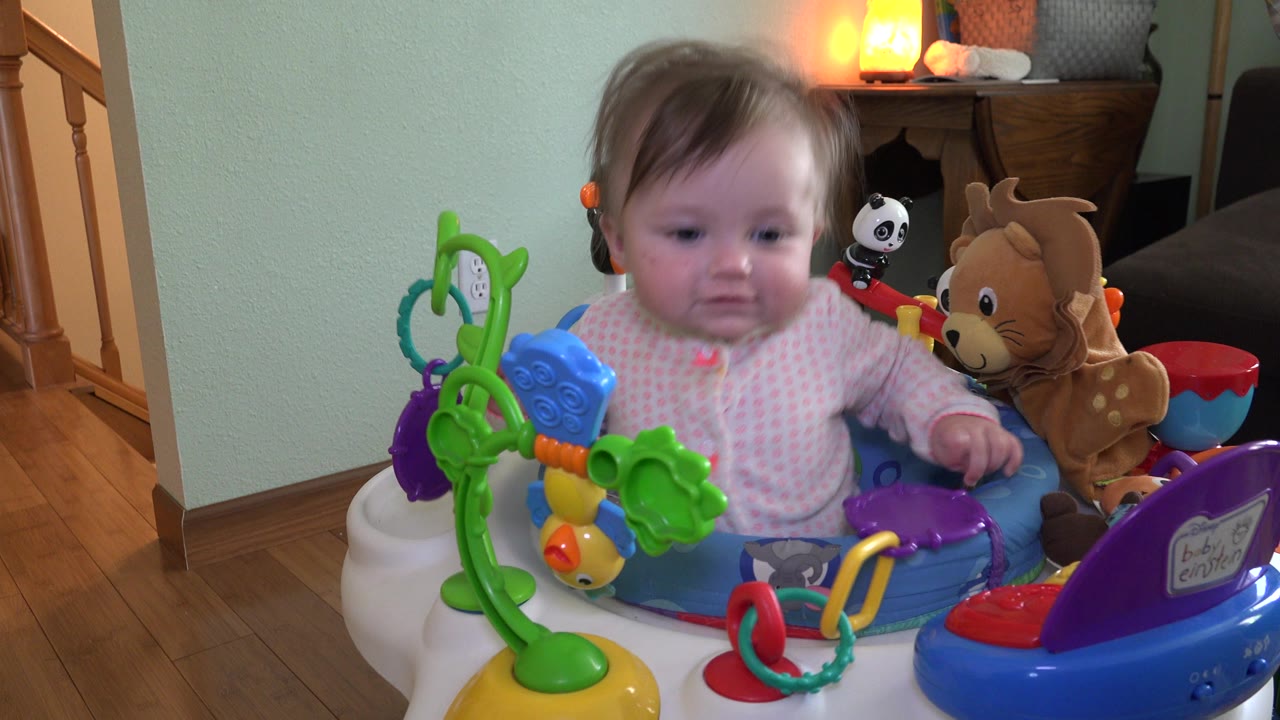 The baby remains seated in the activity seat, looking down toward the toys while a staircase, lamp, and stuffed animals sit behind them in Westminster, Colorado.