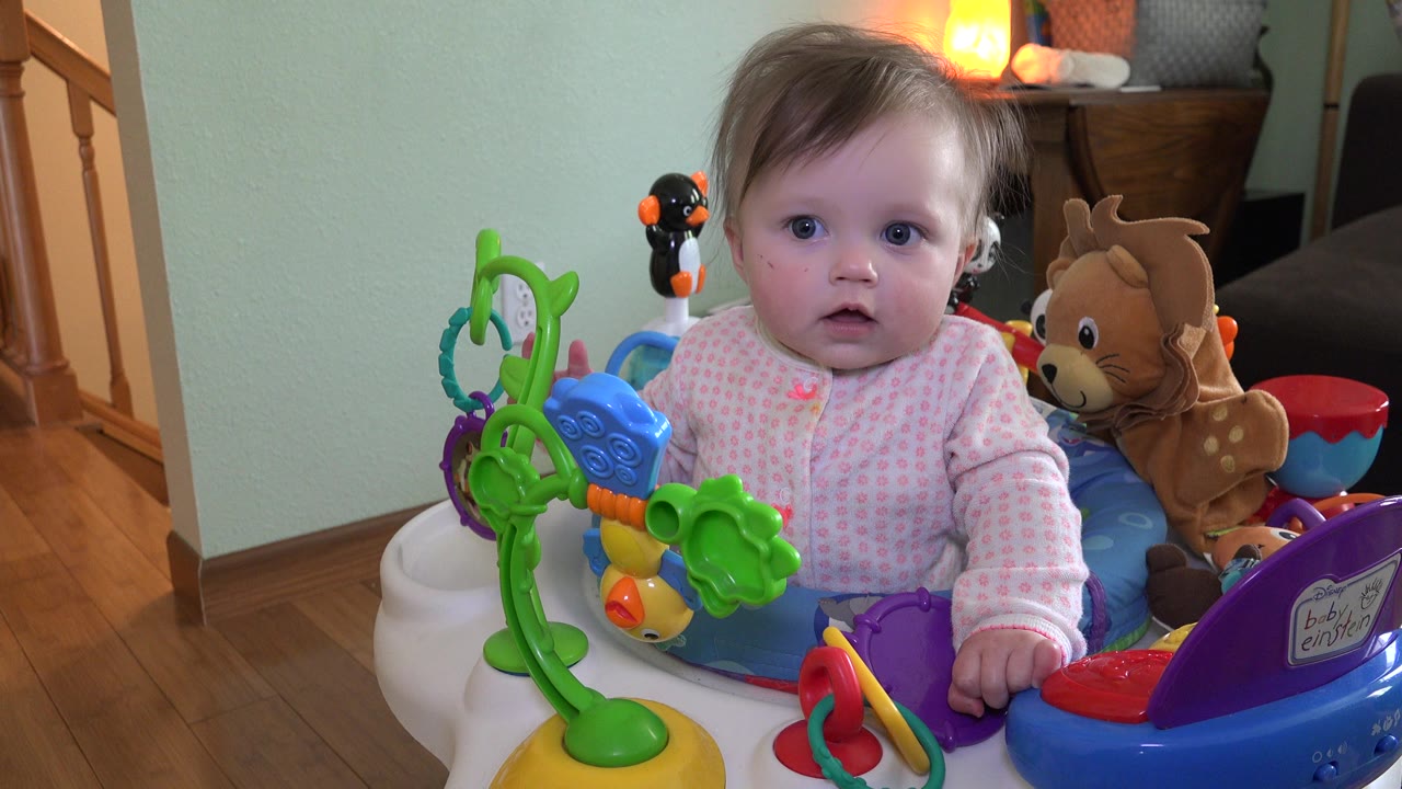 Baby in a pink outfit sitting in a colorful activity chair indoors.
