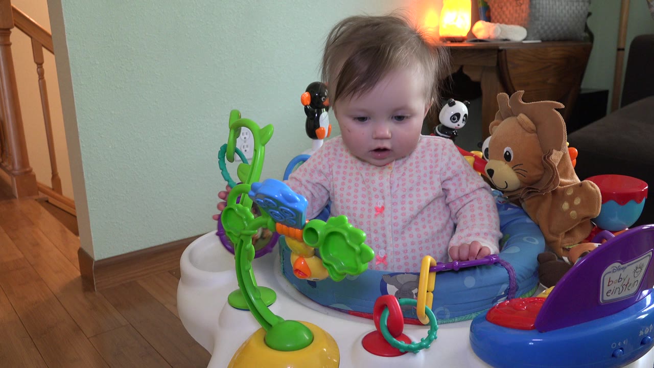 Baby seated in a colorful activity chair with toys in a home interior.