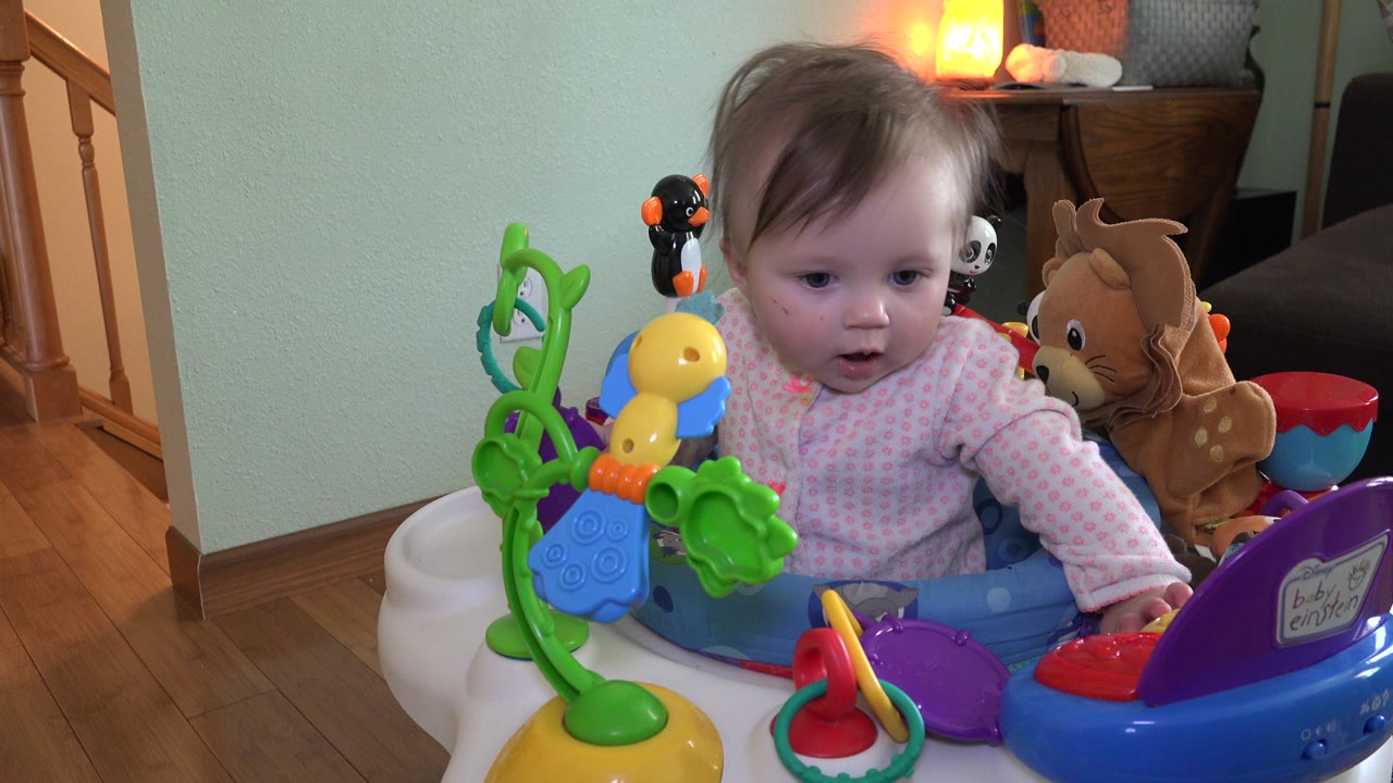 At 0:01, the baby leans forward in the activity chair, looking toward the toys while the wood floor, stairs, stuffed animals, and warm back light remain visible.