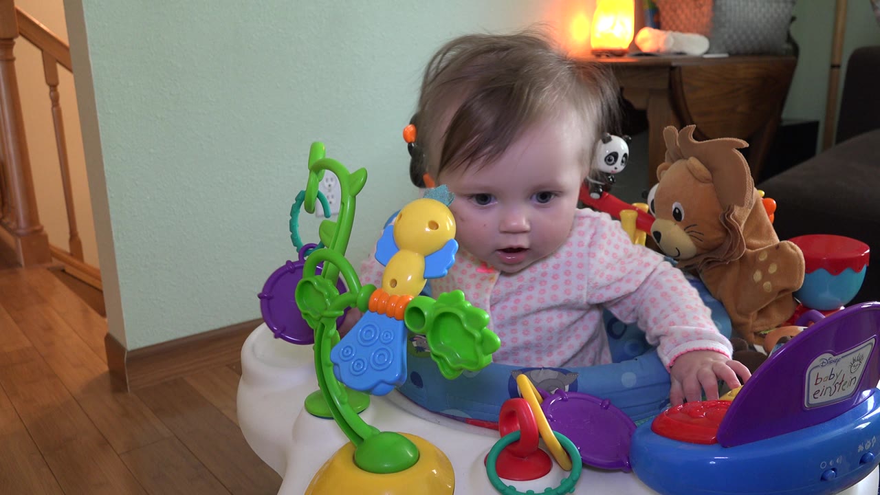 At 0:02, the baby reaches toward the right side of the activity chair, with a green toy structure in the foreground and a stuffed deer behind the seat.