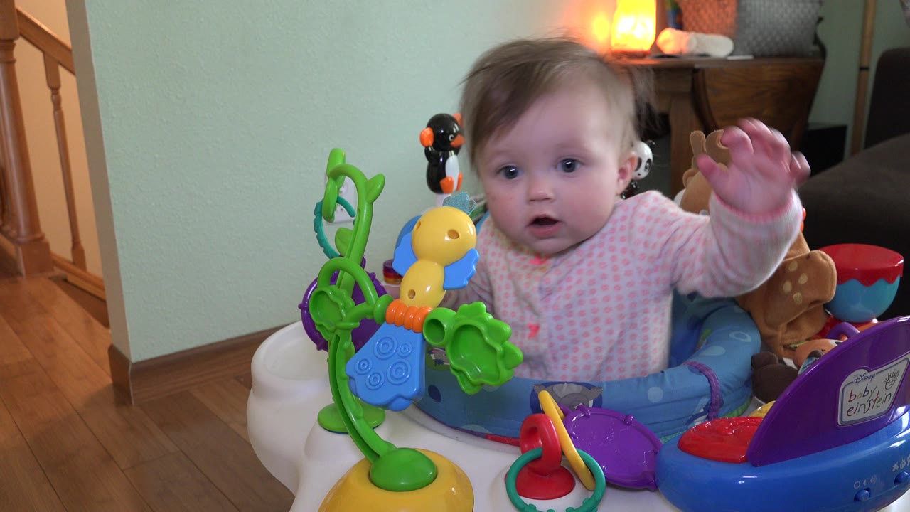 The baby raises one hand from the activity walker as bright plastic toys fill the foreground in the Westminster, Colorado interior.