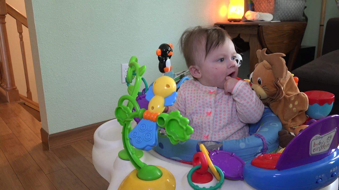 Baby in an activity chair with colorful toys and fingers in mouth.