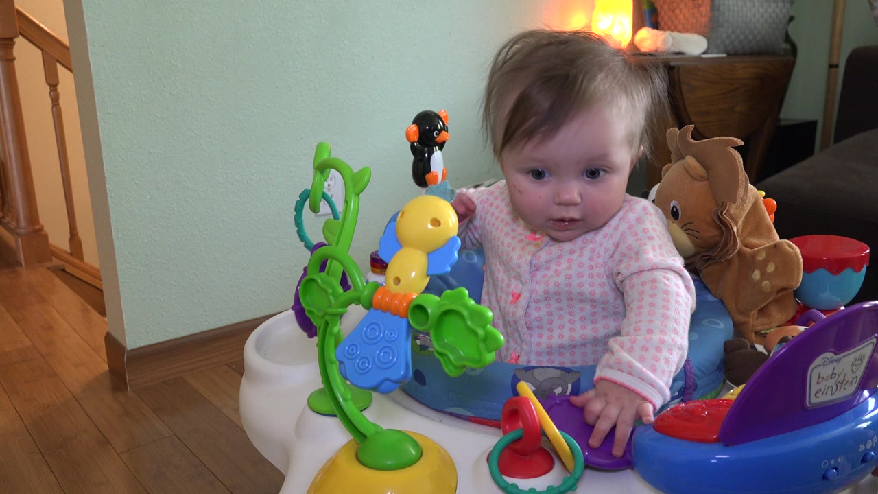 At 0:00, a baby in Westminster, Colorado sits in a blue activity saucer with bright toys in front and a wooden stair rail visible at the left.