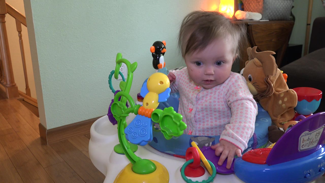 At 0:01, the baby remains centered in the saucer, looking forward past the green and yellow toys with the pale wall and lamp behind.