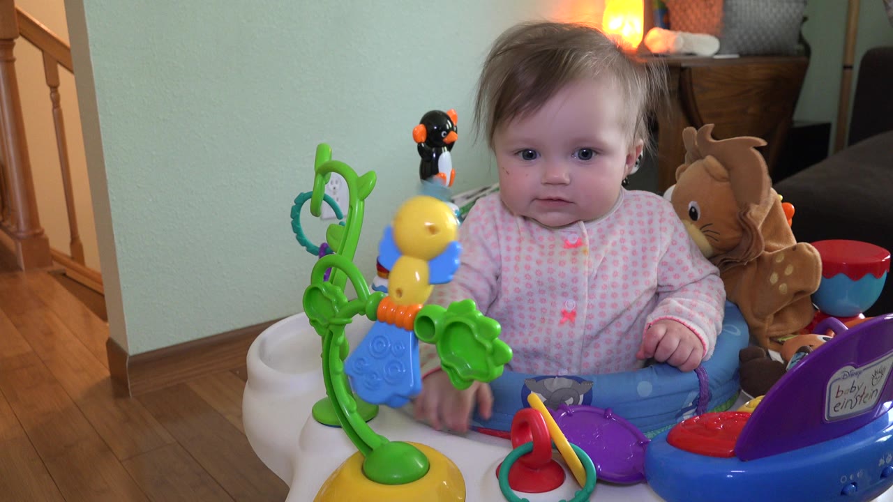 Baby in an activity chair facing colorful toys indoors.