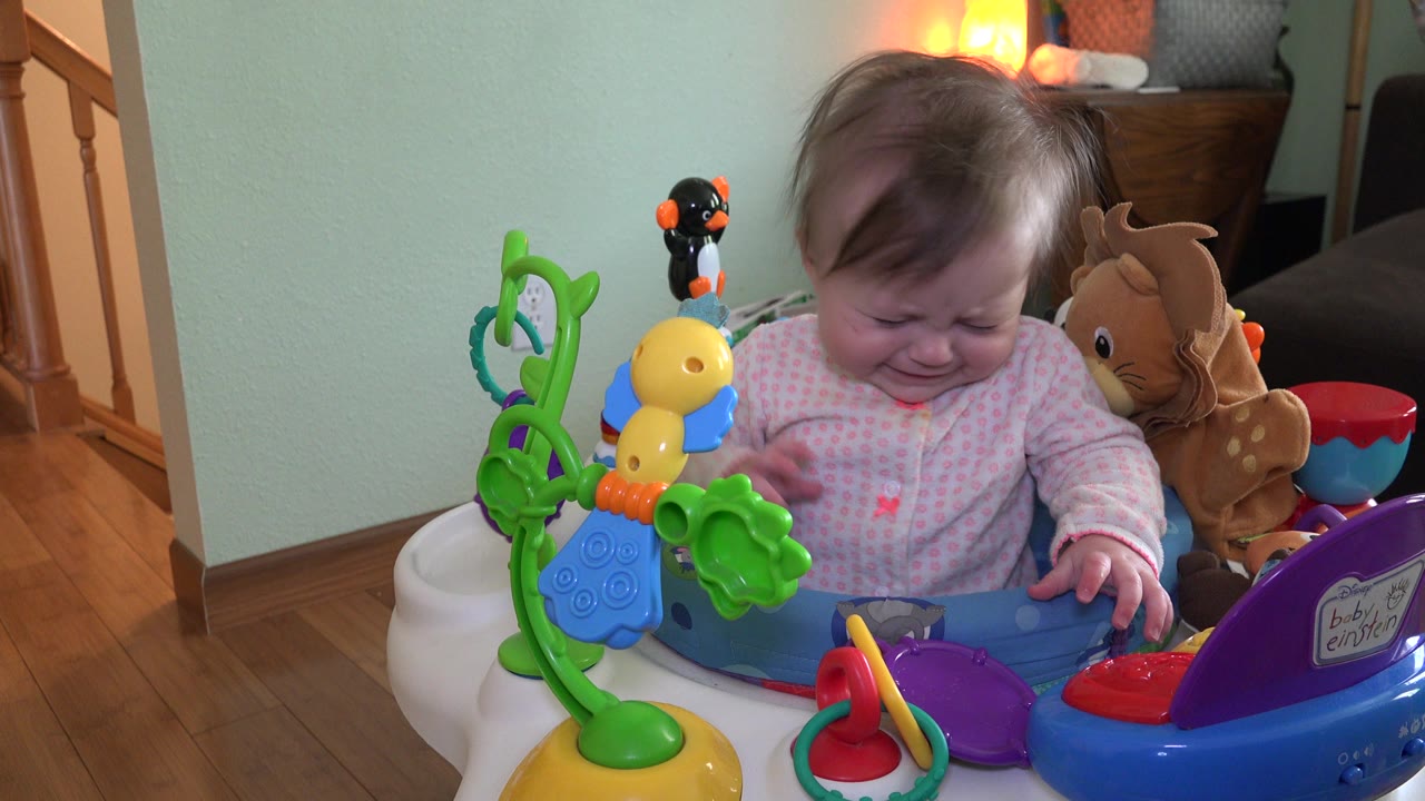 Crying toddler seated in a colorful activity bouncer indoors.
