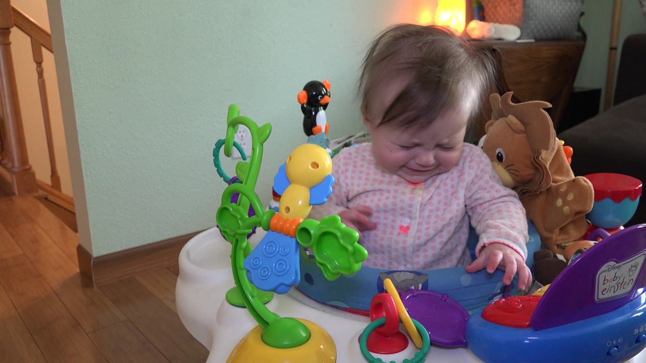 At 0:01, the toddler remains upset in the Westminster, Colorado bouncer, looking down toward the tray with a plush toy tucked near her right shoulder.