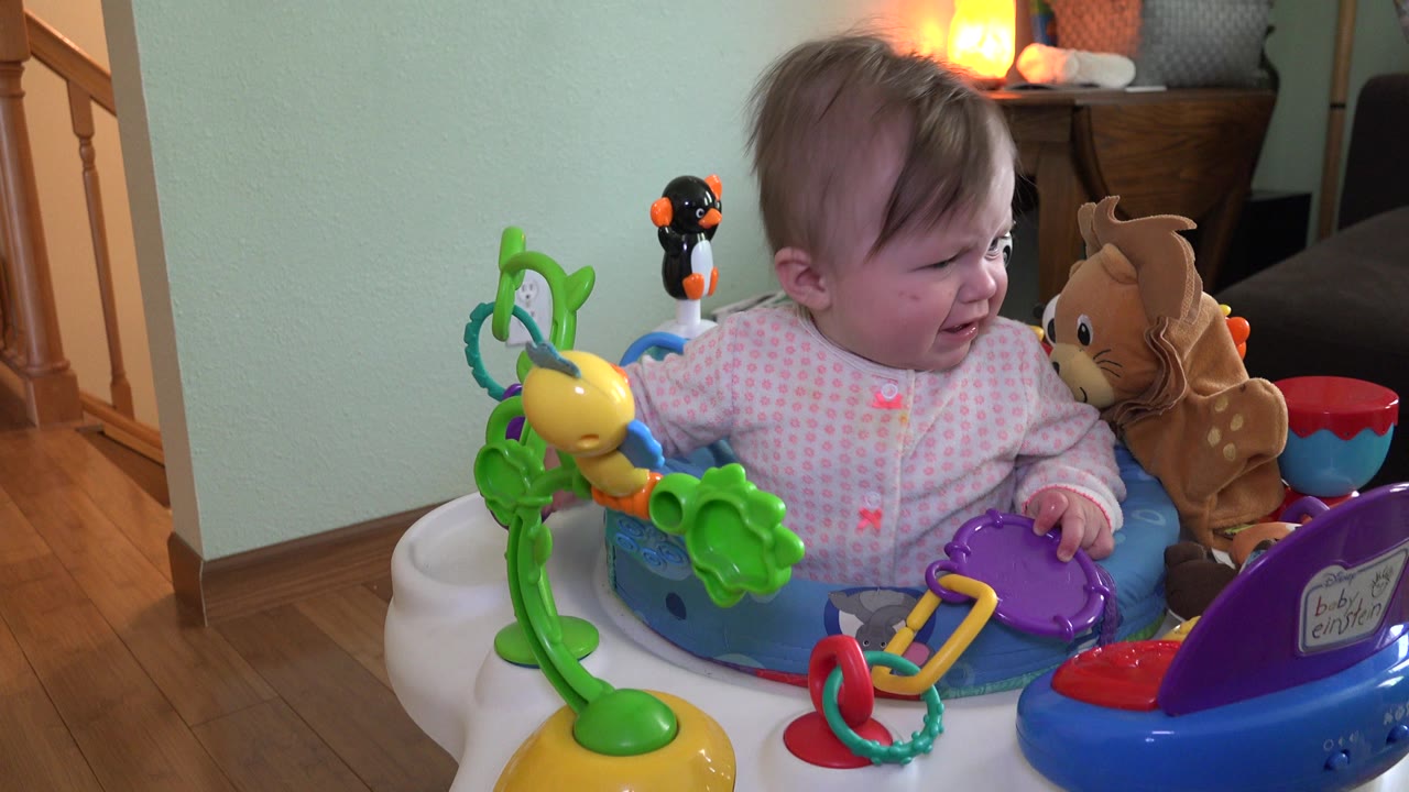 A baby girl in Westminster, Colorado sits in a colorful activity seat, looking toward a plush animal beside her as green and purple toys fill the foreground.