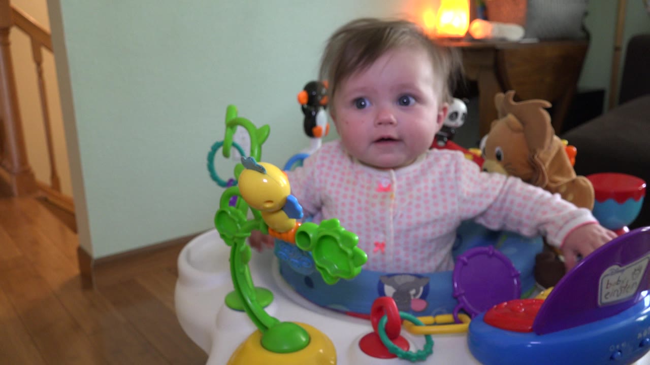 At 0:00, a toddler in a pink patterned shirt sits centered in a blue activity seat with bright toys in an indoor Westminster, Colorado home setting.