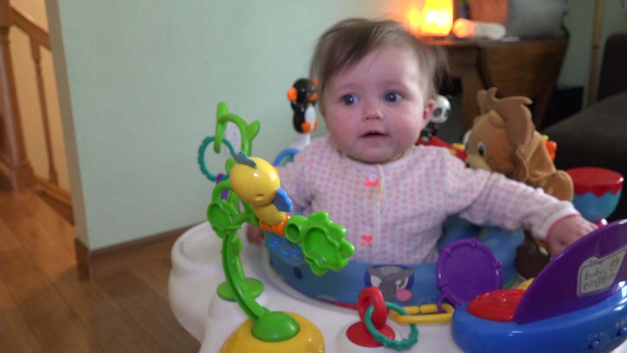 At 0:01, the toddler remains facing forward in the activity seat, with a green toy arm and yellow toy prominent in the foreground and warm room light behind.