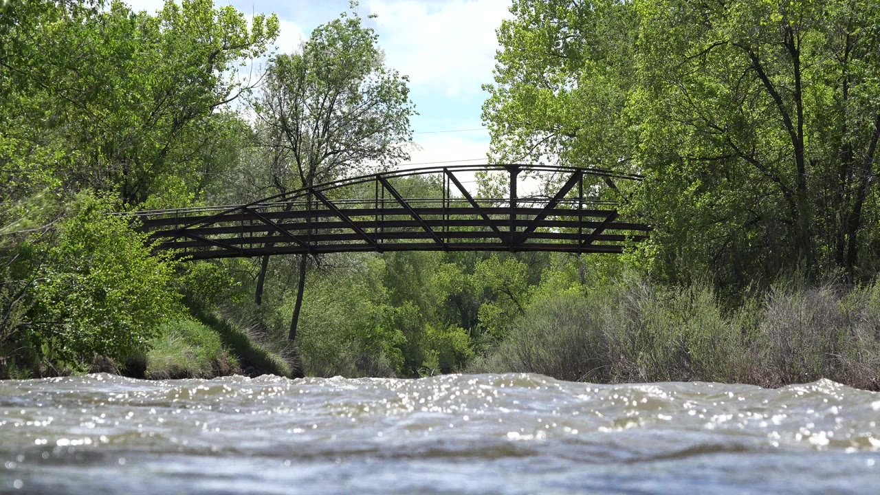 At 0:01 in Westminster, Colorado, the old metal bridge remains centered above choppy sunlit water, with leafy trees and brush behind it and no visible people.