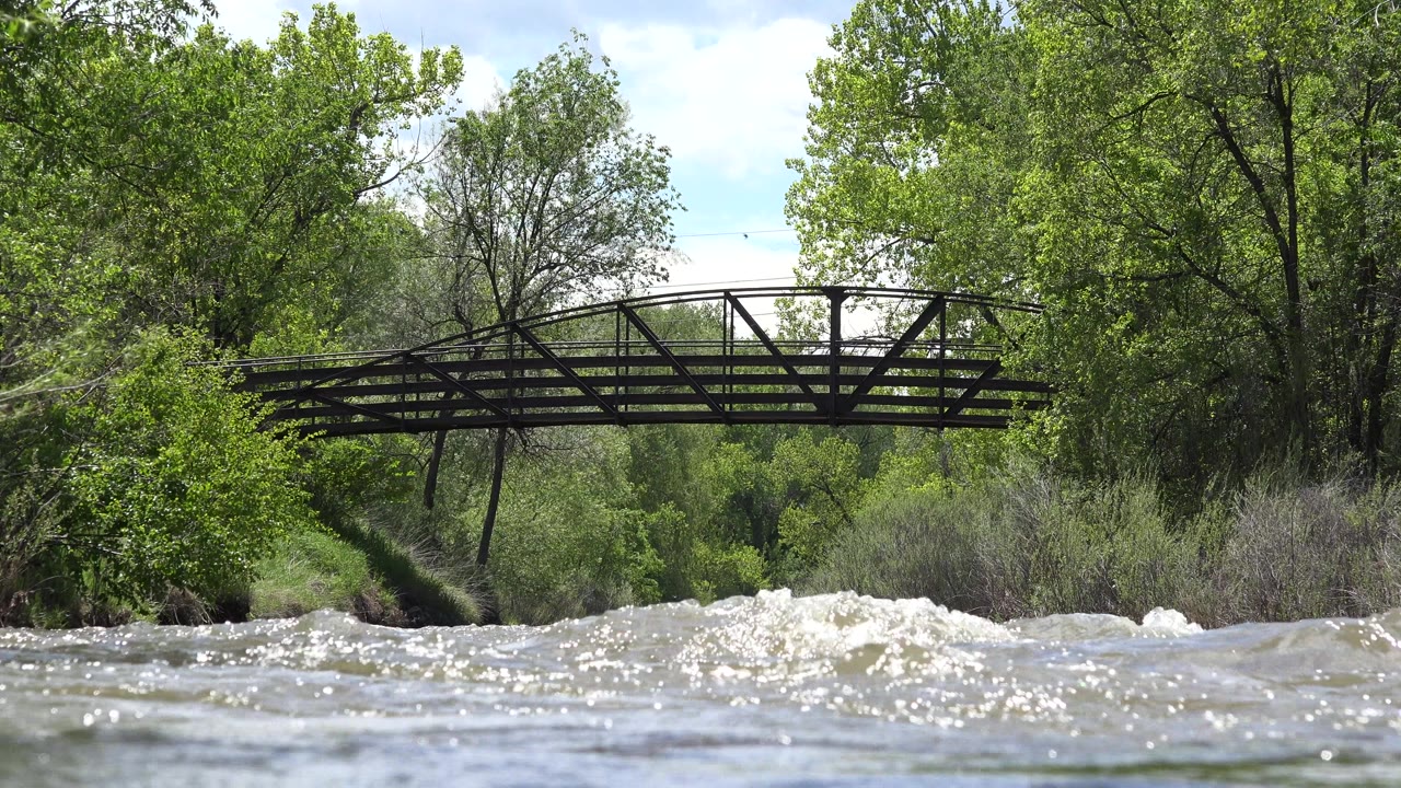 At 0:00 in Westminster, Colorado, a dark pedestrian bridge spans choppy sunlit water with green trees behind it and no visible people.