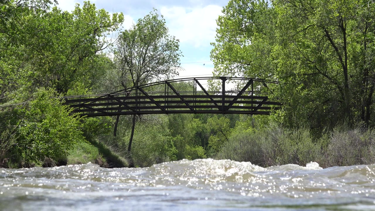 At 0:01 in Westminster, Colorado, bright ripples fill more of the foreground beneath the bridge while the tree line remains dense and empty of people.