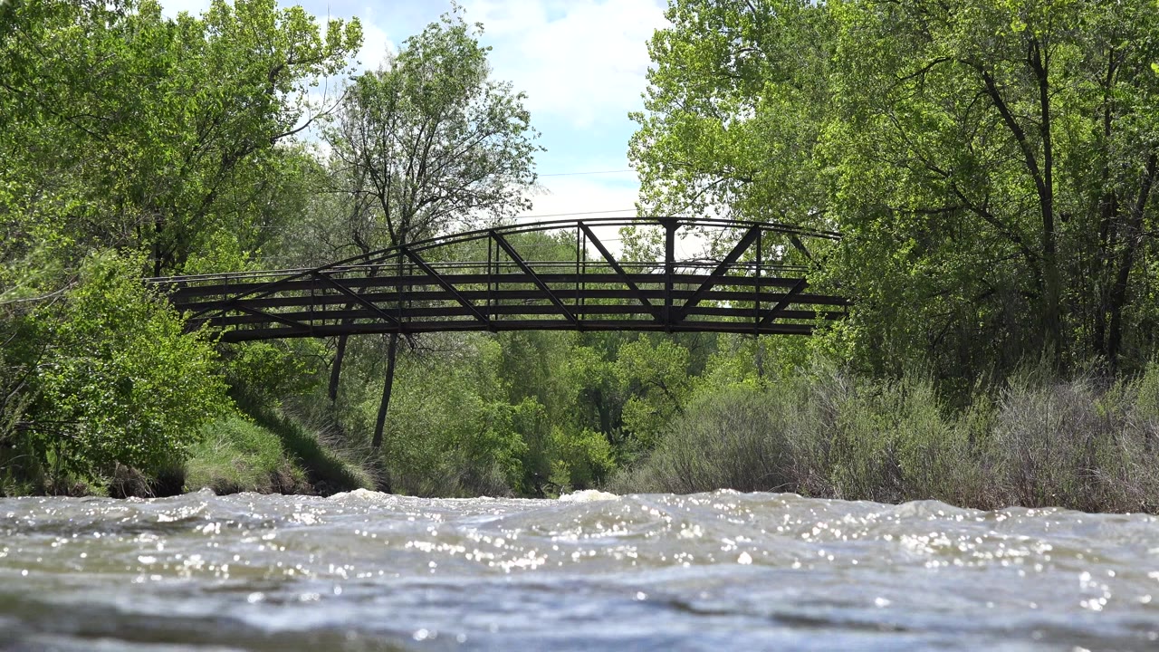 River waves glint in the foreground while the pedestrian bridge and leafy banks remain centered in the Westminster, Colorado frame, with no visible people.
