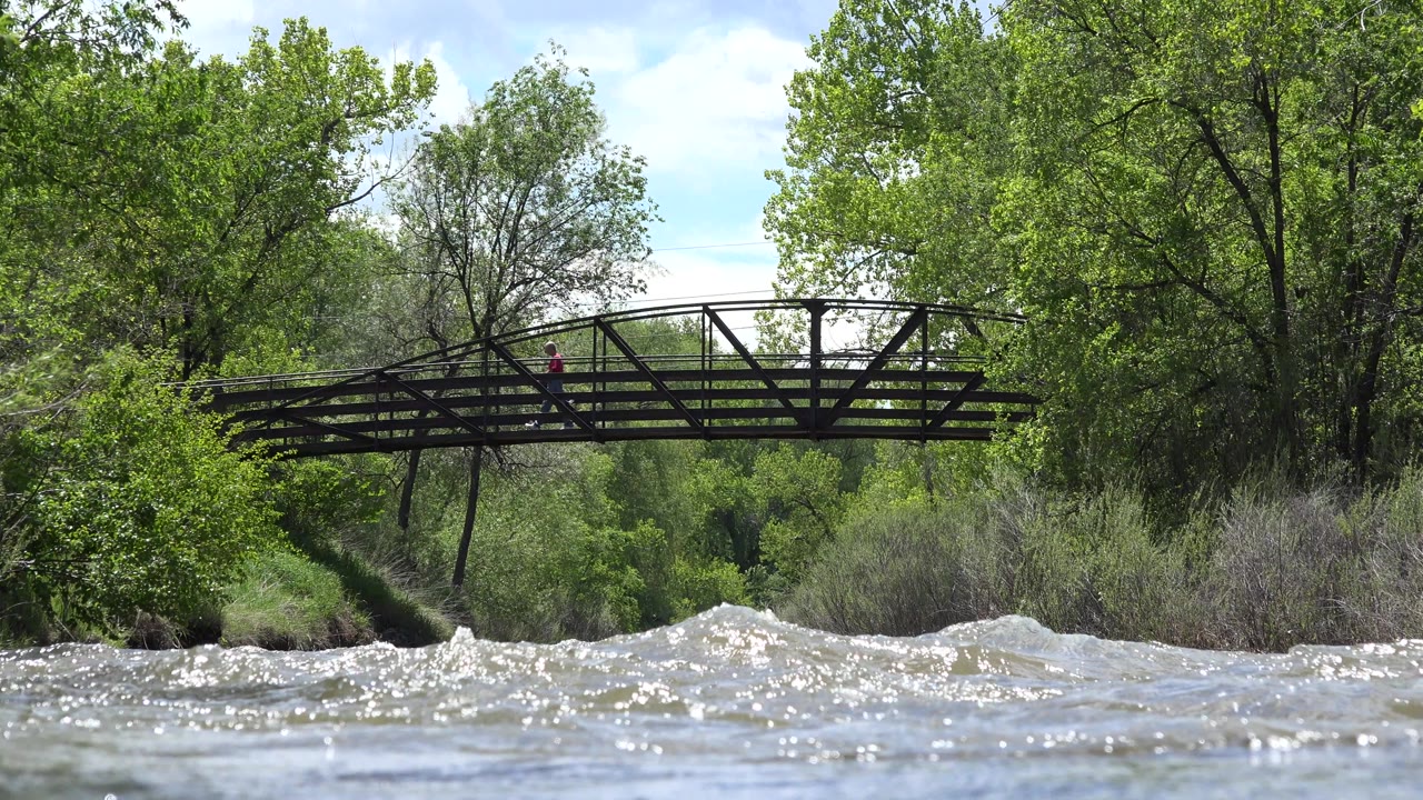 Rushing river water below a metal footbridge with trees behind it.