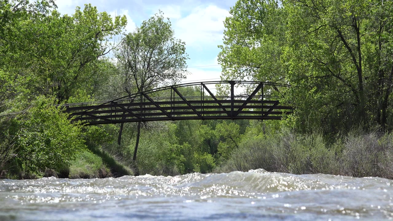 At 0:00 in Westminster, Colorado, bright rushing river water crosses the foreground below a dark pedestrian bridge, with dense green trees behind it and no visible people.
