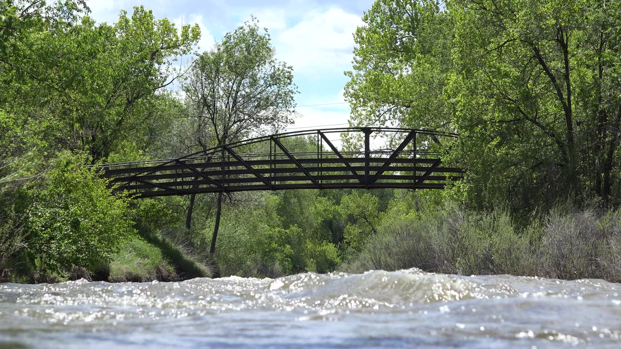 At 0:01 in Westminster, Colorado, foamy whitewater rises higher across the foreground while the bridge remains centered against green riverside vegetation, with no visible people.