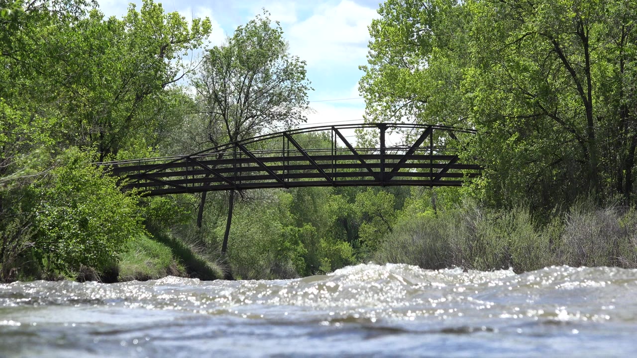 Bright ripples rise in the foreground while a bridge spans the wooded riverbank in Westminster, Colorado, with no visible people.