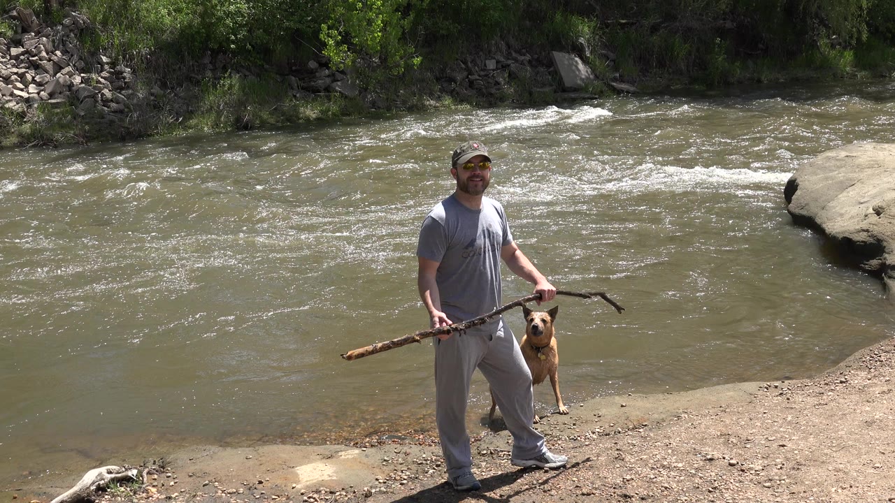 In Westminster, Colorado, a man in gray clothing holds a long stick beside a tan dog on a dirt bank while rippled water moves behind them.