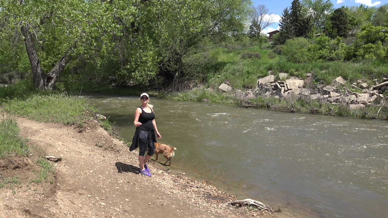 Woman in a white cap and sunglasses stands with a brown dog on a sunlit dirt bank beside a flowing river in Westminster, Colorado.