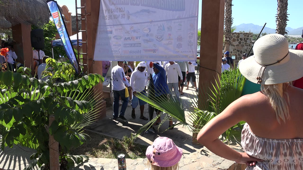 Crowded shaded beachside walkway with plants and people under a sign.