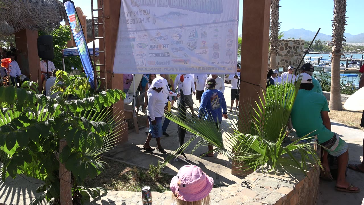 In Playita, Baja California Sur, a woman in a wide sun hat stands close to the camera while people move beneath a hanging sign, with plants in front and boats and water visible in the background.