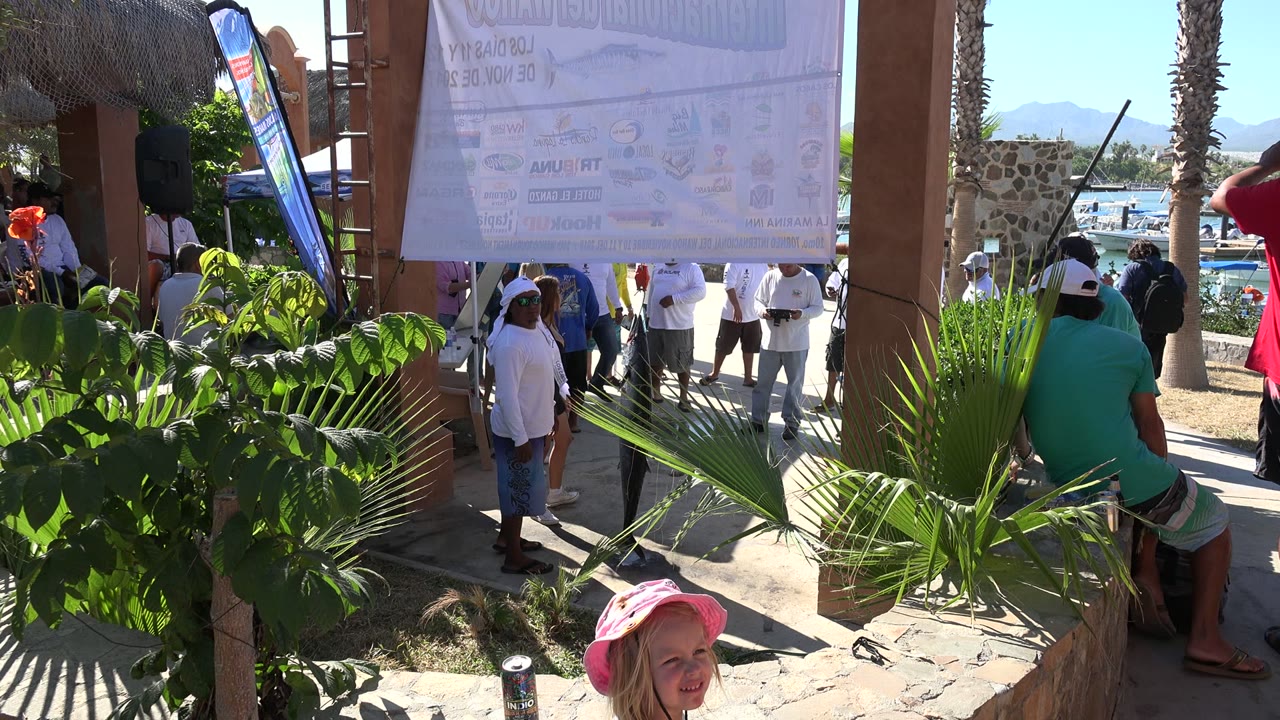 Crowd outside under a large hanging sign near palms and water.