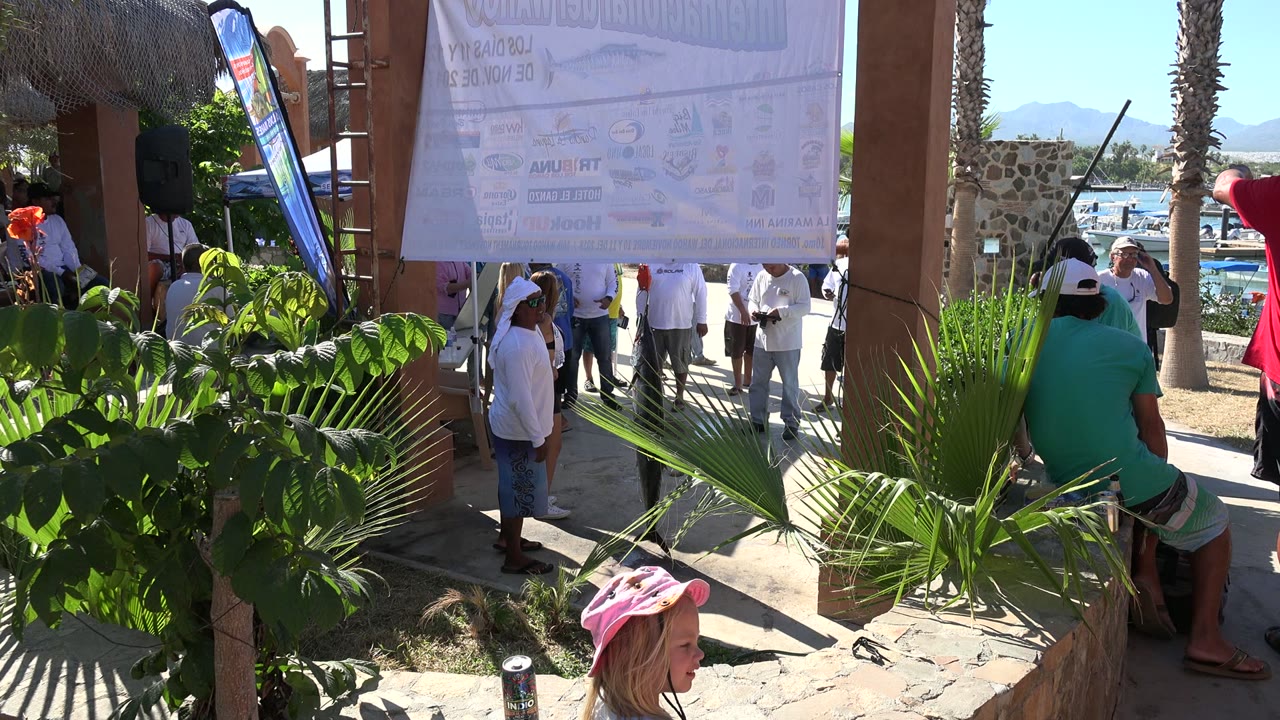 At 0:01 in Playita, Baja California Sur, the crowd remains under the sign as a seated person in a green shirt faces away and boats appear through the opening.