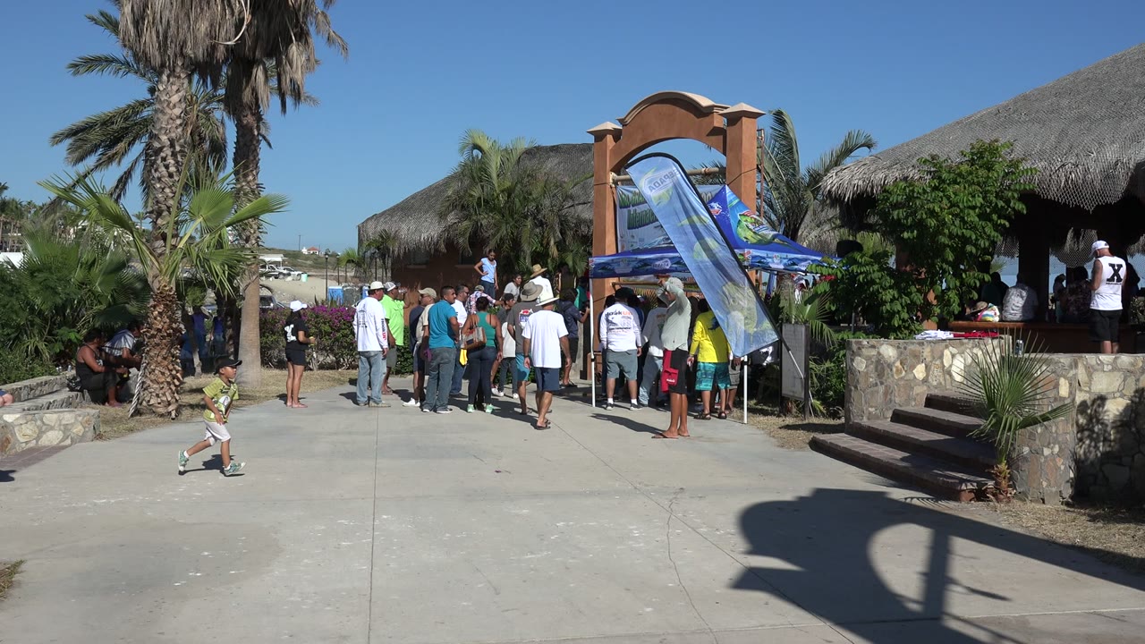 In Playita, Baja California Sur, the child moves farther right across the open pavement while adults remain grouped beside the flags and shaded kiosk.