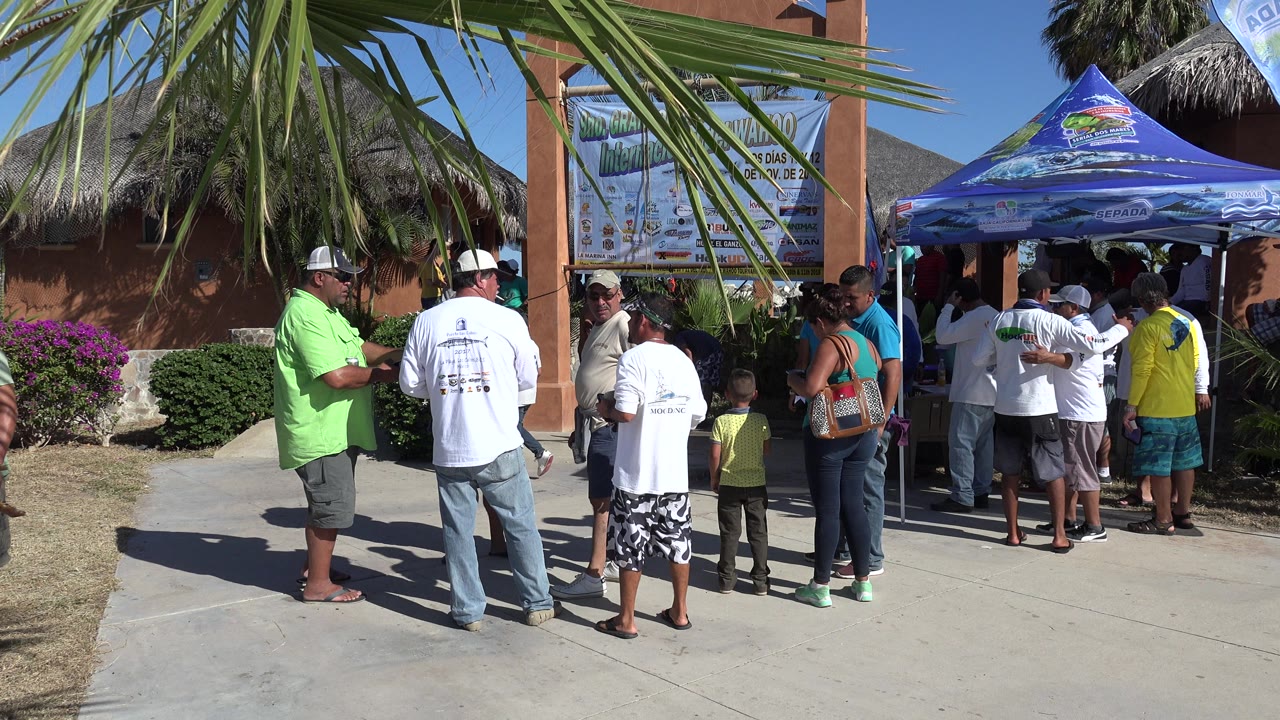 A small group waits beside the booth in Playita, with a man in a bright green shirt at left, white-shirted visitors in the center, and a child near the entrance.