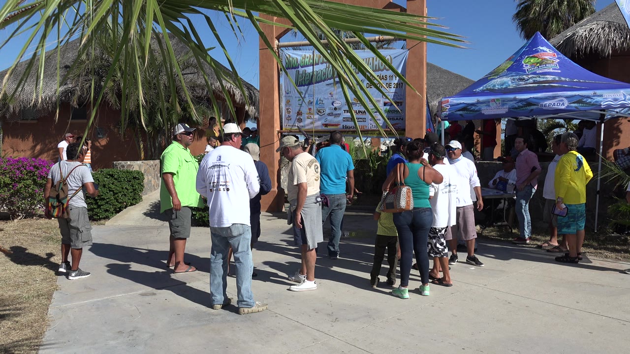 People stand and talk on a bright walkway in Playita, Baja California Sur, Mexico, with palm fronds overhead, a large entrance sign, and a blue canopy at the right.